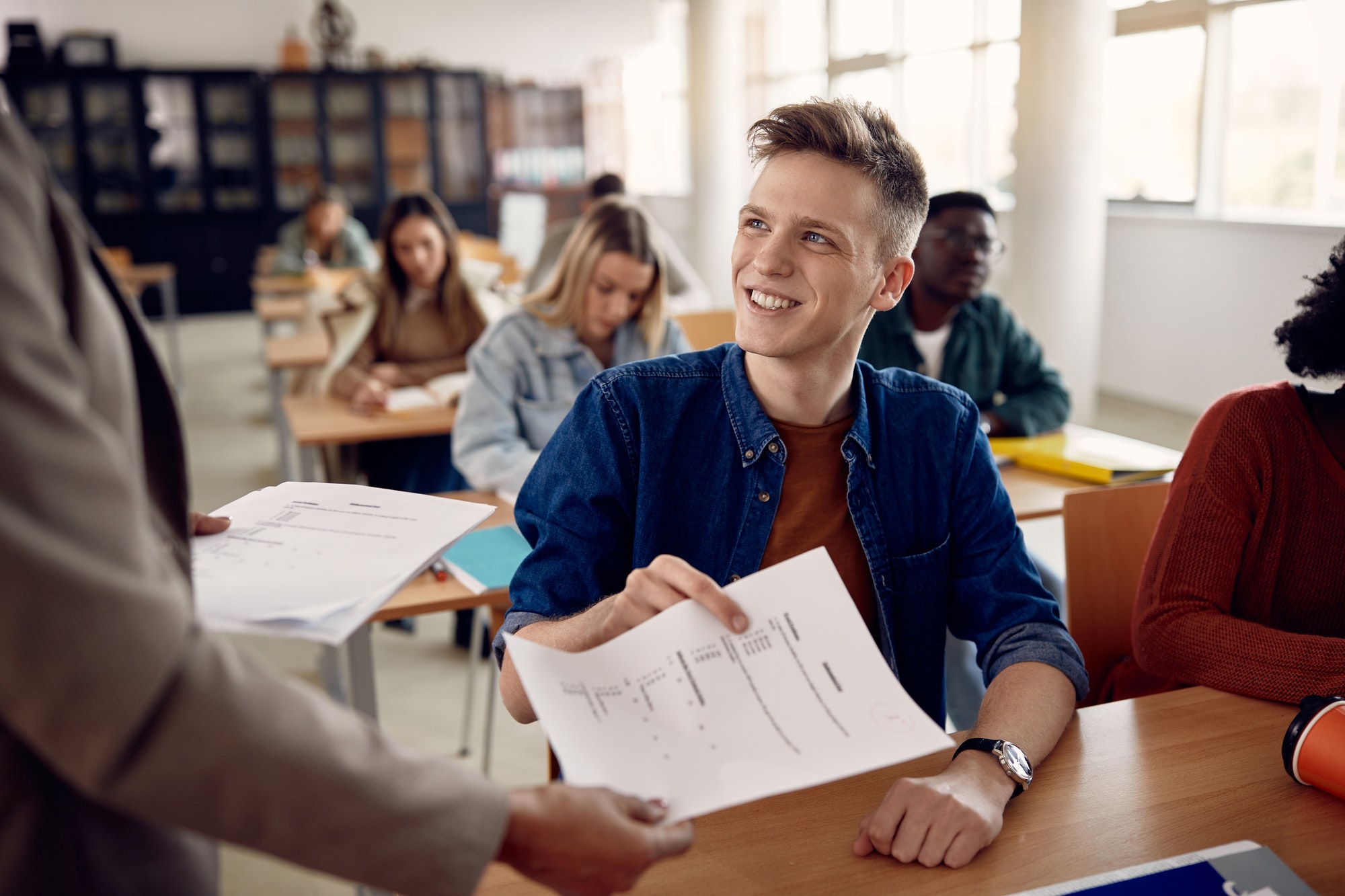 Happy college student taking exam paper from a teacher in the classroom.